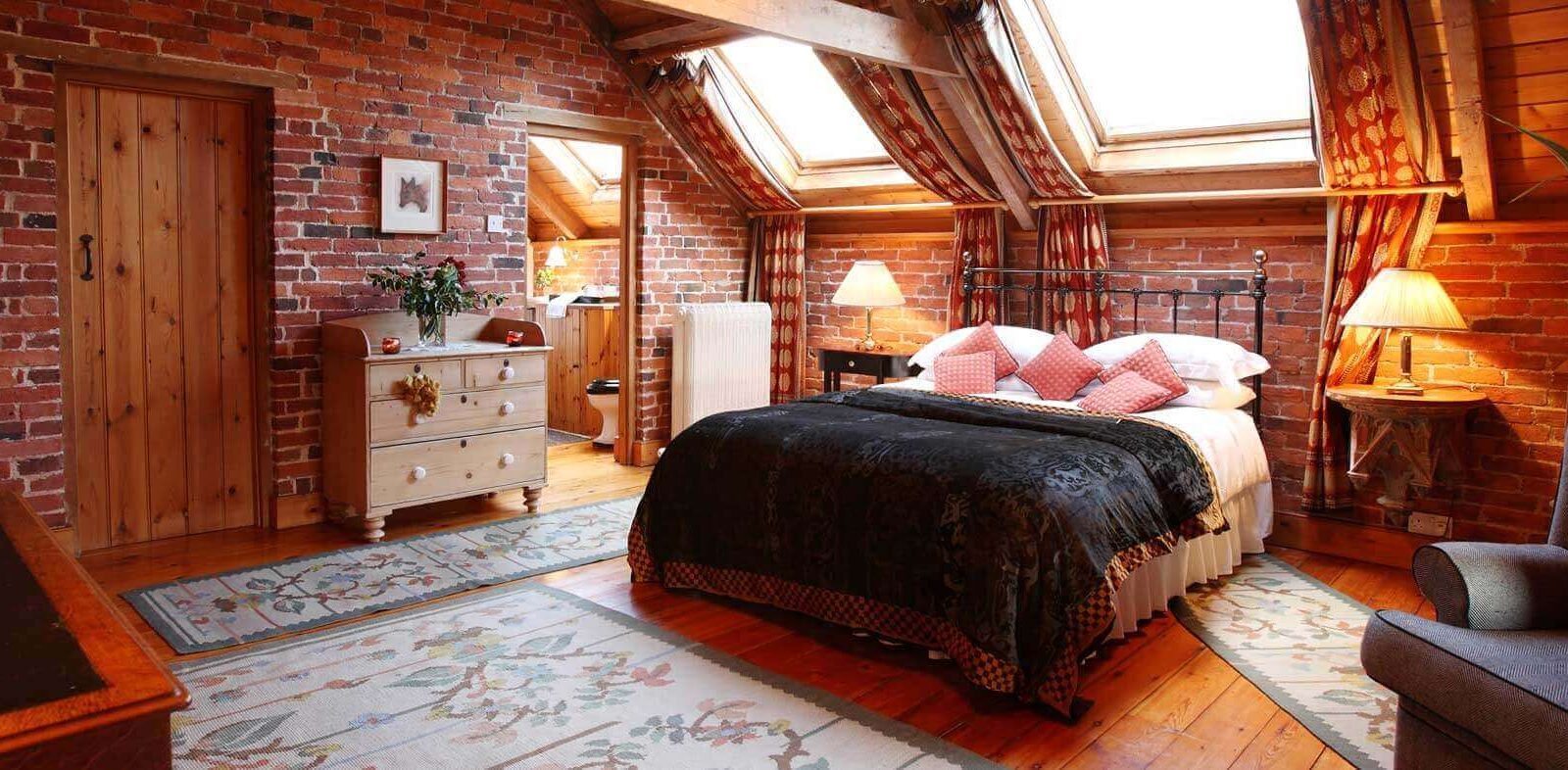 A cozy bedroom with a rustic design reminiscent of a haybarn, featuring exposed brick walls and wooden ceiling beams. The room is furnished with a double bed, an armchair, a wooden dresser, and a rug. Skylights above the bed let in natural light, creating the perfect farm stay vibe at Pennard Hill Farm.