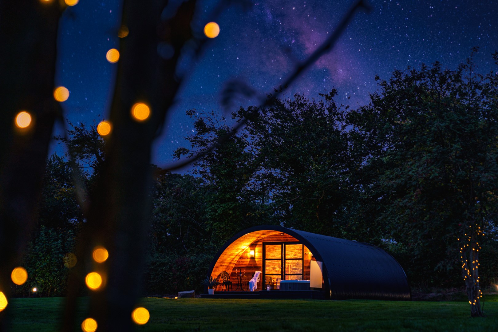 A cosy, curved cabin with lit interiors sits in a dark, wooded area under a starry dark night sky. Warm lights on nearby trees add a magical touch to the serene setting—perfect for Winter Getaways UK.