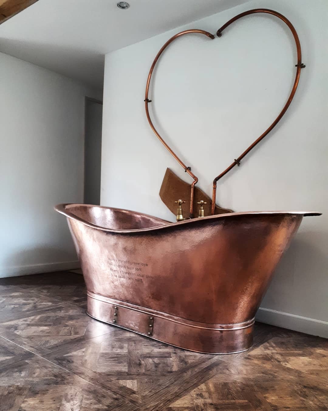 A luxurious, freestanding copper bathtub with a high back sits on a herringbone-pattern wooden floor. Above the tub, two copper pipes form a large heart shape on the white wall, adding artistic flair to the minimalist bathroom— perfect for romantic winter getaways in the UK.