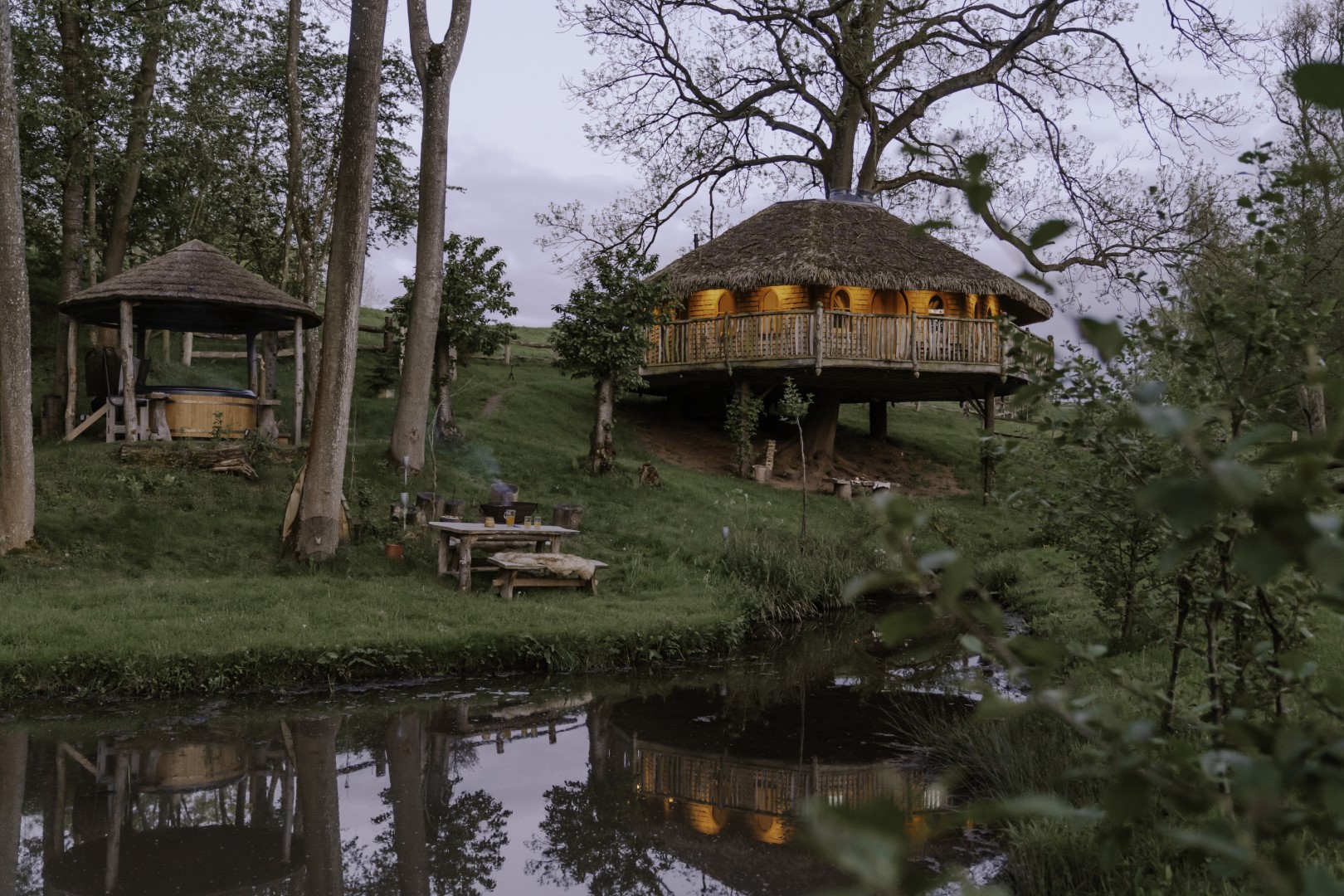 A rustic wooden treehouse with a thatched roof is nestled among trees near a tranquil pond, offering the perfect Winter Getaways UK experience. The treehouse glows warmly from within as evening falls. A cosy outdoor seating area with log benches and a small gazebo is set up beside the water.