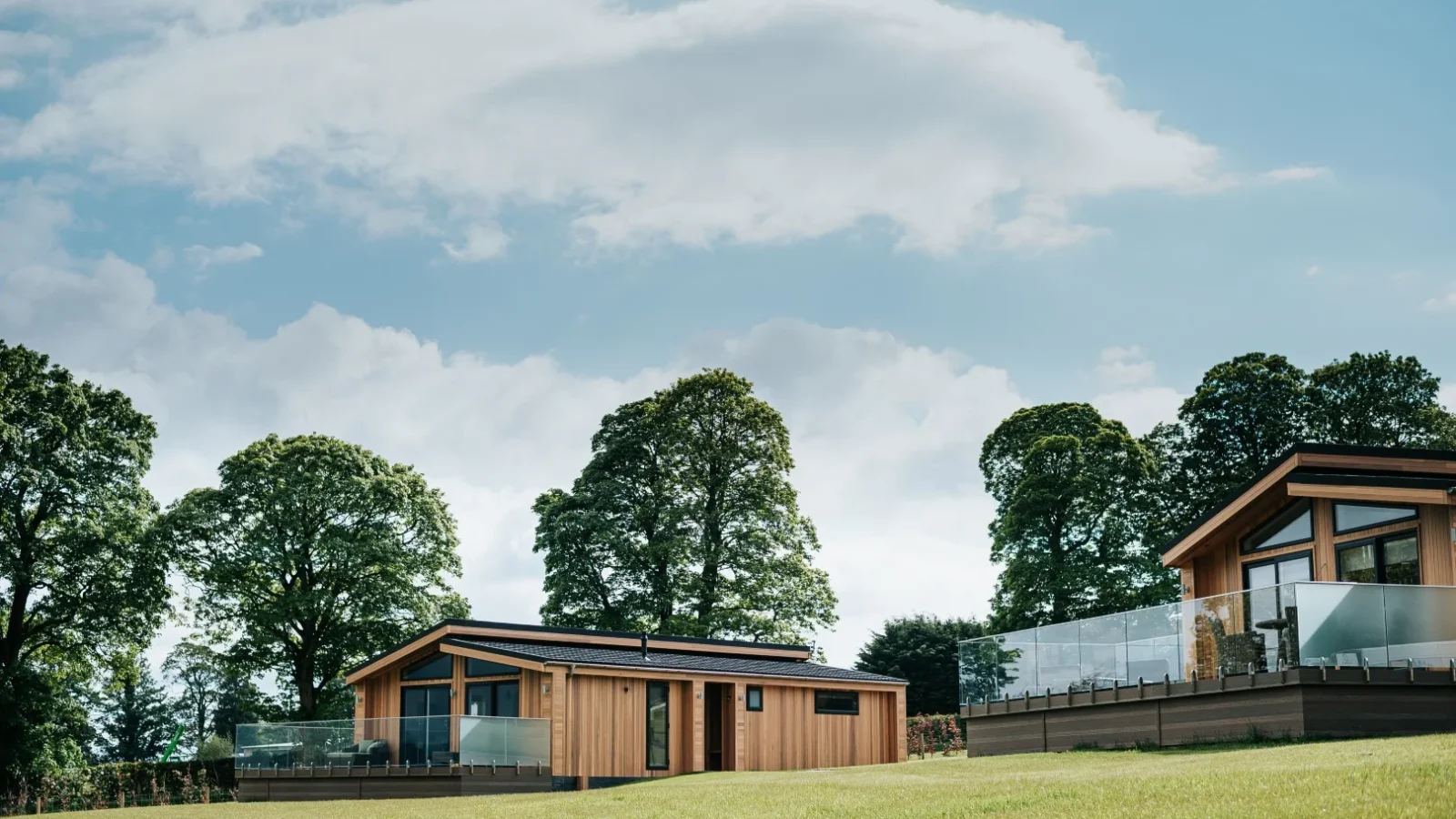 A scenic view of modern wooden cabins, known as Bowland Retreat Lodges, situated on a grassy hillside. The cabins feature large windows and glass railings, offering unobstructed views of the lush green trees surrounding the area. The sky is clear with a few clouds.