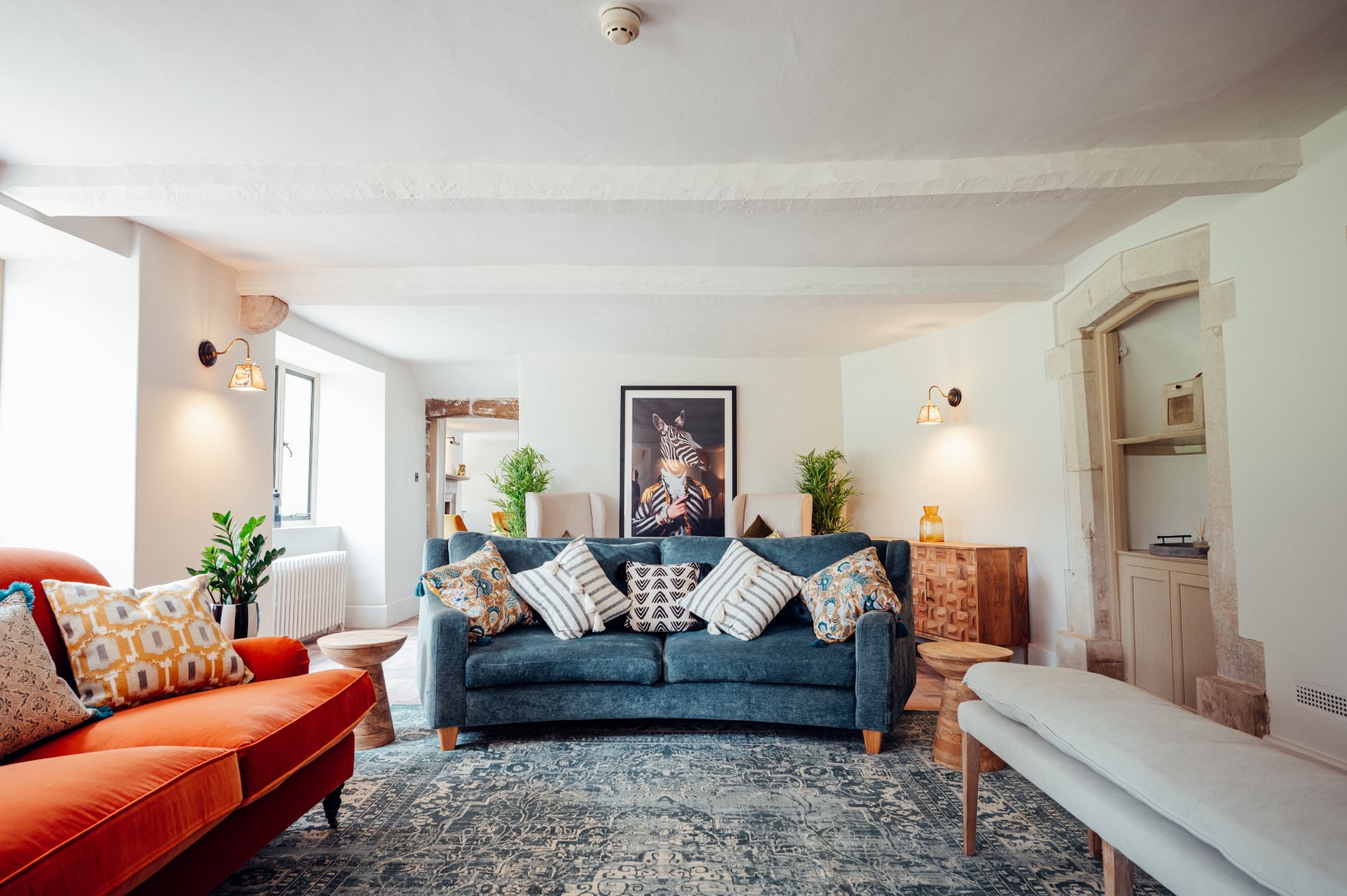 A cosy living room with a blue sofa, an orange sofa, and a white bench. The sofas have assorted patterned cushions. A framed artwork is on the wall behind the blue sofa. The room has white walls, wooden furniture, plants, and a large patterned rug. Dog friendly accommodation near to dog friendly beaches in the UK.