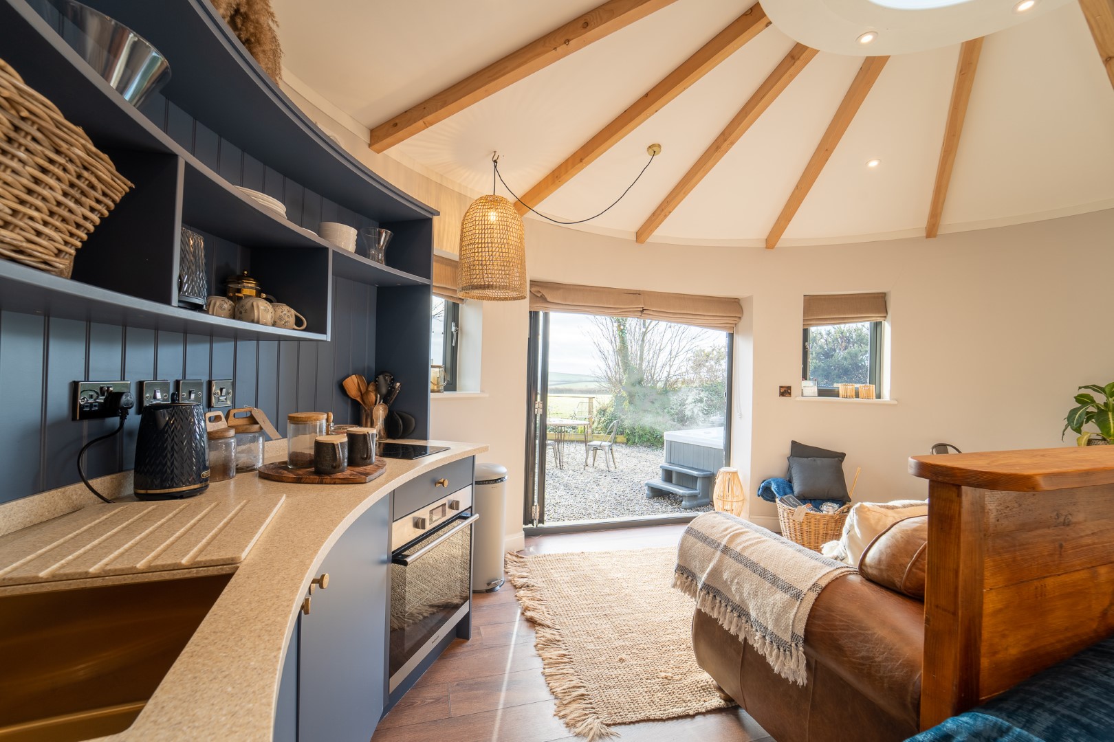 A cosy, modern yurt with a curved kitchen area featuring wooden countertops, blue cabinets, and various kitchen appliances. The interior includes a sofa, woven baskets, a woven light fixture, and open double doors leading to an outdoor view of greenery and gravel. Nearby dog-friendly beaches add to the appeal for pet lovers.