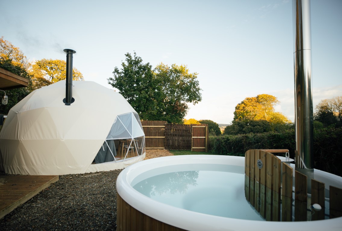 A geodesic dome tent sits nestled in a leafy setting, with trees in the background under a clear sky. In the foreground, there's a round outdoor hot tub with a wooden enclosure. The tranquil and inviting scene is perfect for nature lovers and close to dog-friendly beaches. Ideal for a dog friendly holiday in the UK. 