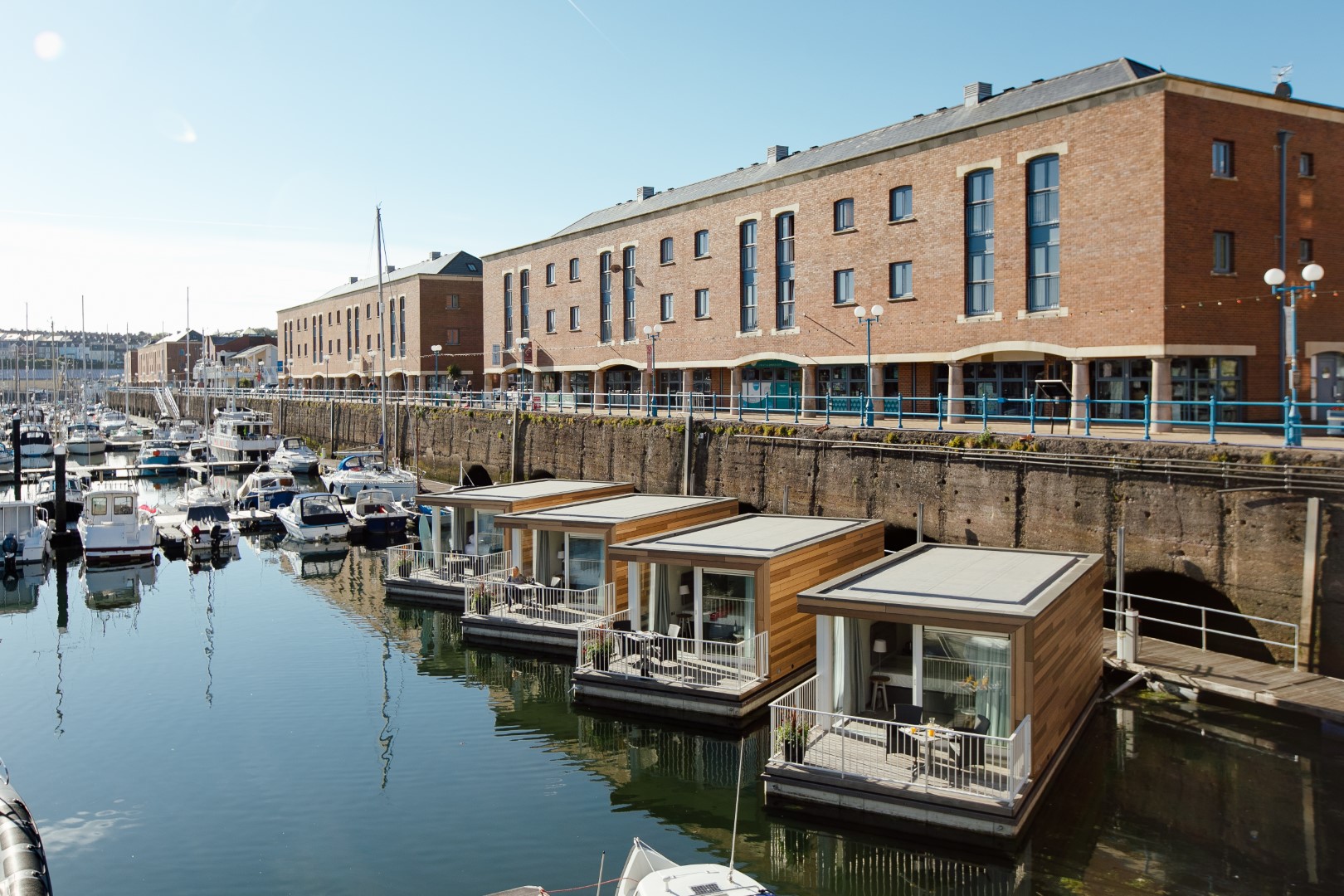 A waterfront scene shows modern floating homes on a calm body of water. Behind the homes, a brick building with multiple windows and a walkway is visible. Several boats are docked in the marina to the left, near dog-friendly beaches. The sky is clear and sunny.