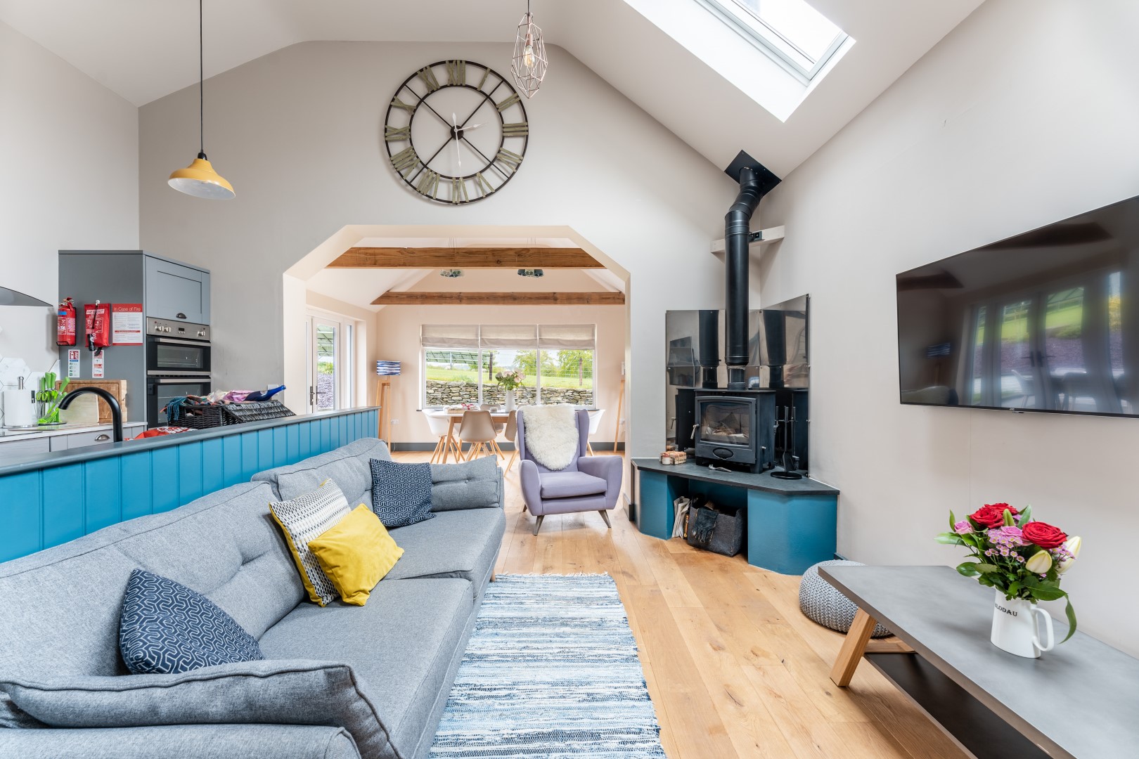 A modern, airy living room with a high ceiling and skylight. It features a grey sectional sofa, colorful cushions, a flat-screen TV, a wood-burning stove with a chimney, and a large clock on the wall. A kitchen and dining area are visible in the background, perfect for unwinding after enjoying dog-friendly beaches nearby.