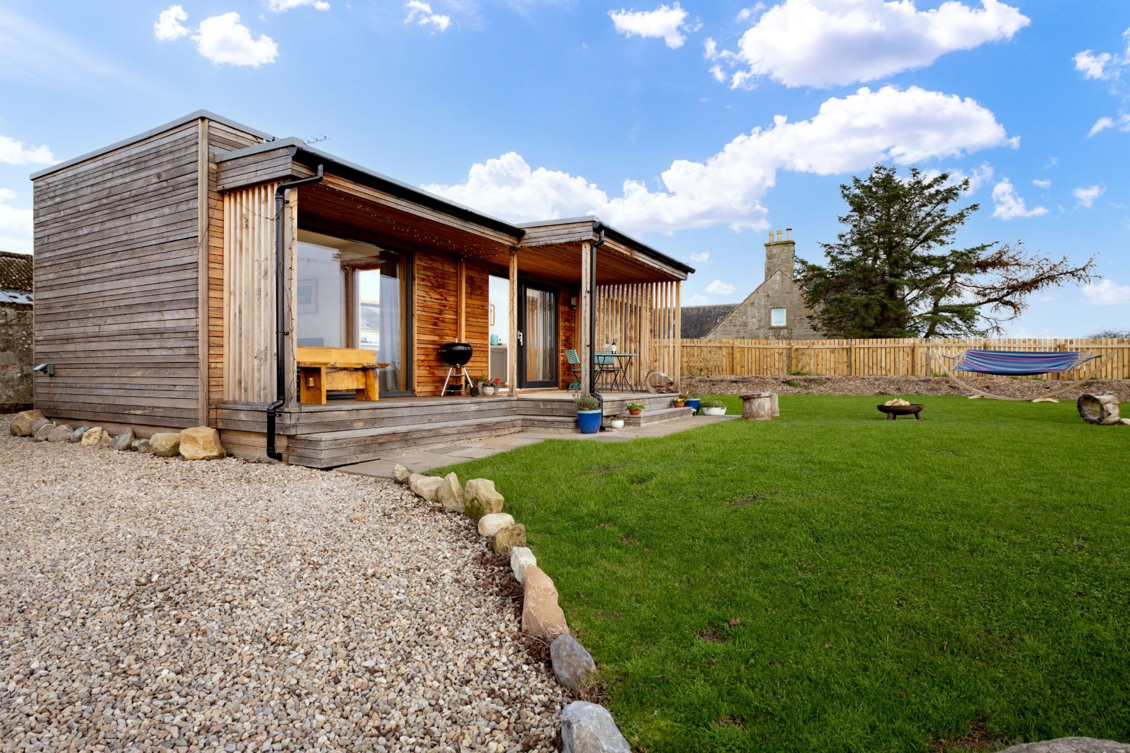 A modern wooden cabin with a deck, situated in a grassy yard with a gravel pathway. The cabin features large glass doors, a hammock in the background, and a small fire pit area on the lawn. Nearby dog-friendly beaches add to the appeal of this clear, pleasant setting. Book a dog friendly coastal stay in the UK