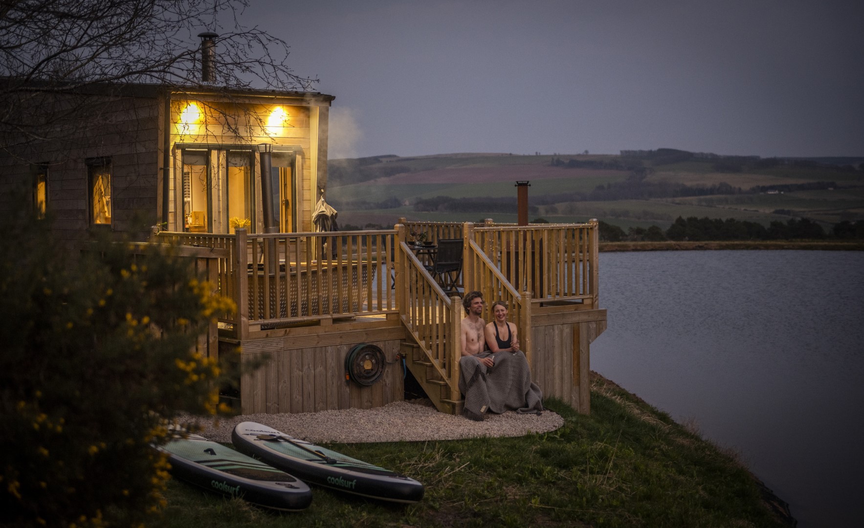 A couple sits wrapped in blankets at the bottom of a wooden staircase leading to a cozy, illuminated cabin on a loch side at dusk. Two paddleboards lie nearby. The landscape features gentle rolling hills and a serene loch under a calm evening sky, perfect for romantic weekend breaks with a hot tub.