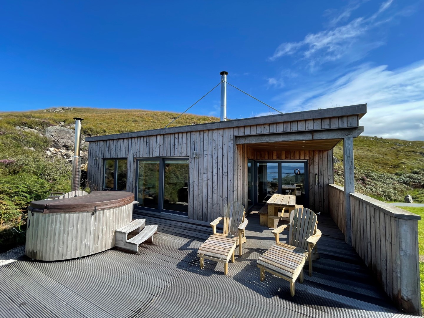 A modern wooden cabin with large glass windows and a deck furnished with Adirondack chairs and a wooden hot tub. Ideal for coastal holidays, the cabin is set against a backdrop of grassy hills under a bright blue sky with a few clouds.