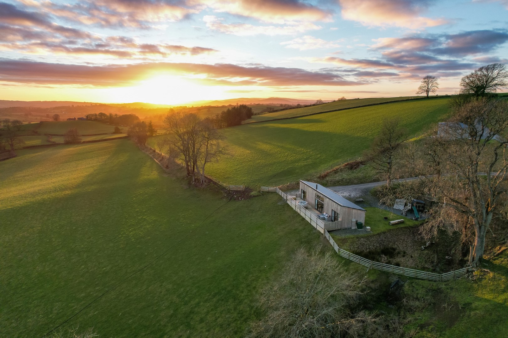 A sunset view of a green hilly landscape with a modern house situated on the right side of the image. The house, ideal for a family getaway in the UK, is surrounded by trees and a fenced yard, with rolling hills and scattered trees stretching into the distance under a sky with scattered clouds.