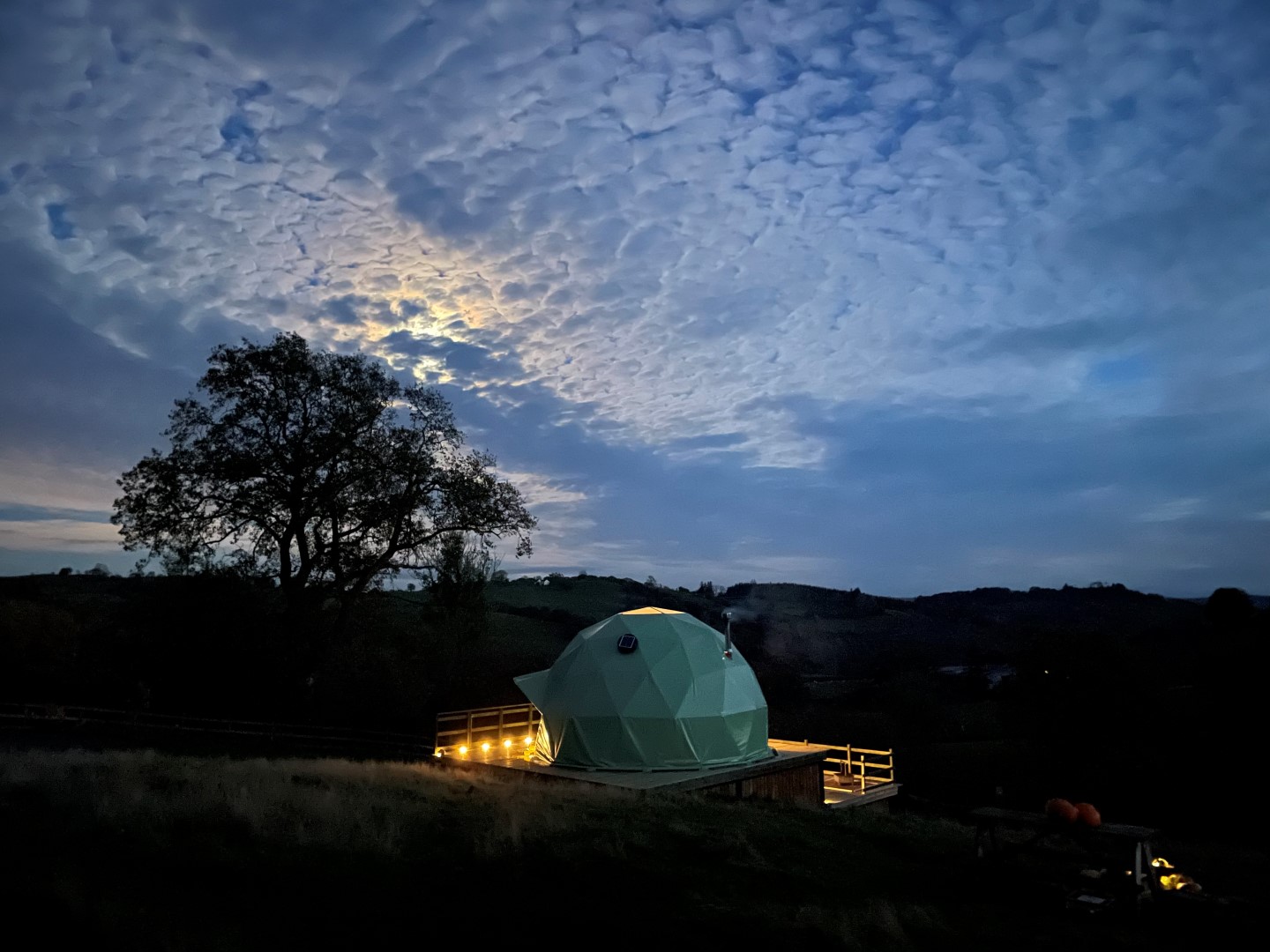 A geodesic dome structure sits illuminated on a wooden deck under a twilight sky with textured, cloud-streaked patterns. A large tree and hilly landscape are visible in the background, creating a serene, natural setting—perfect for winter glamping in the UK.