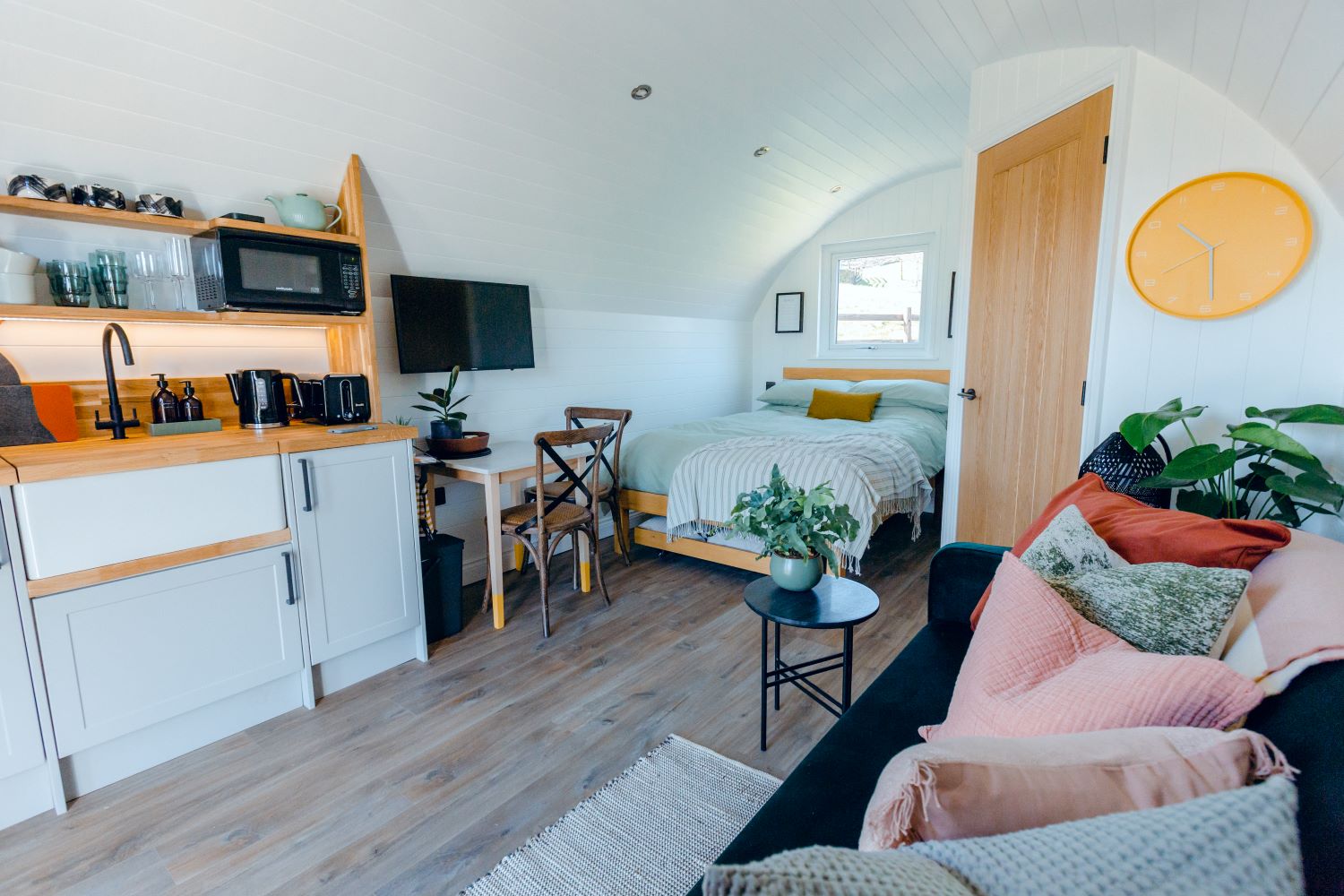 A well-lit, cosy pod perfect for winter glamping in the UK features a kitchenette with wooden countertops, a microwave, and small appliances. A bed is placed near a window, with a wall-mounted TV nearby. A sofa with colourful pillows and a coffee table with plants complete the living space.