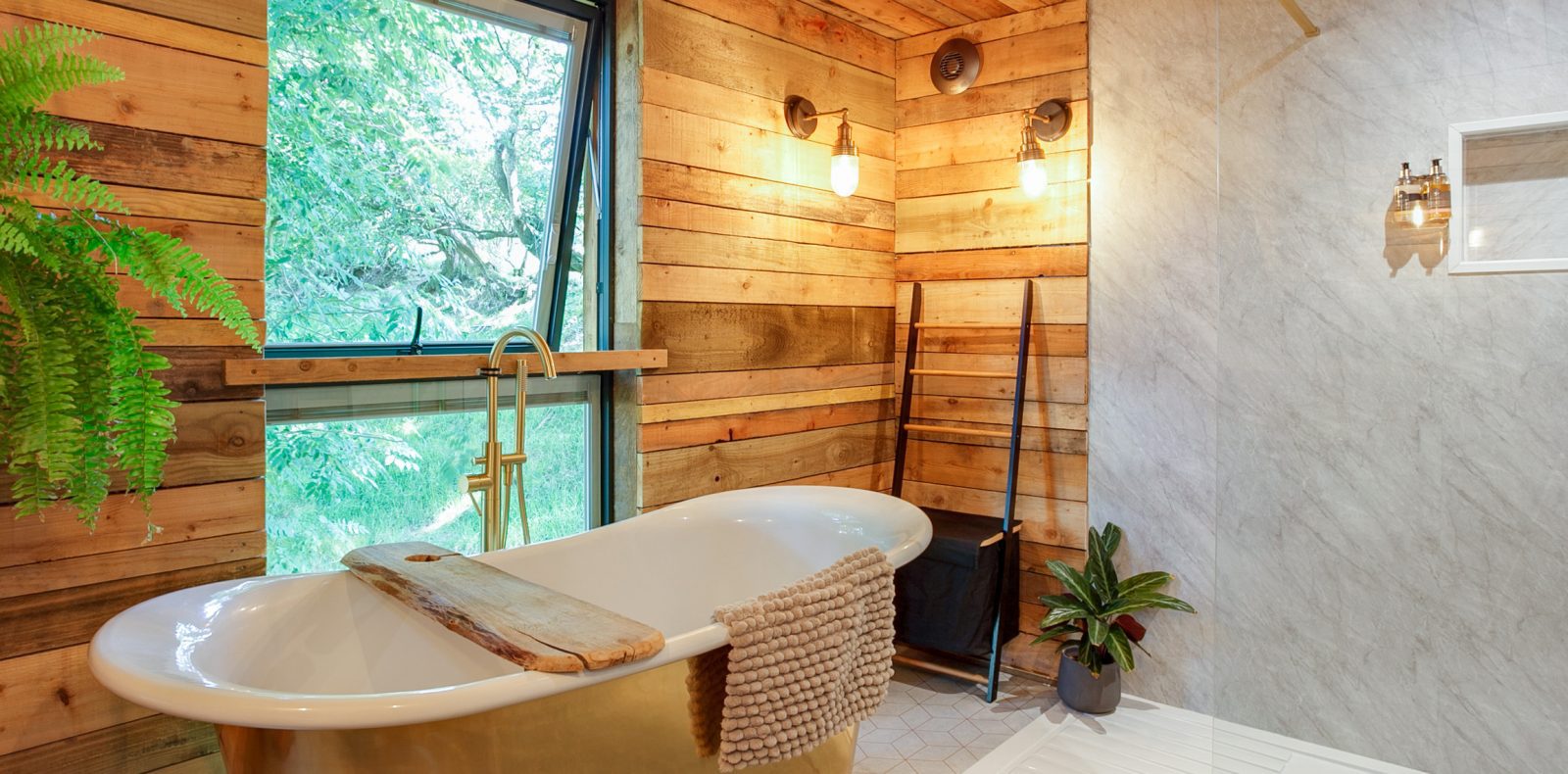 A rustic bathroom in the Prince Llywelyn Treehouse features a freestanding white bathtub with a wooden tray. The room boasts wooden panel walls, a large window with views of greenery, and charming potted plants. Two wall-mounted lights provide warm illumination.