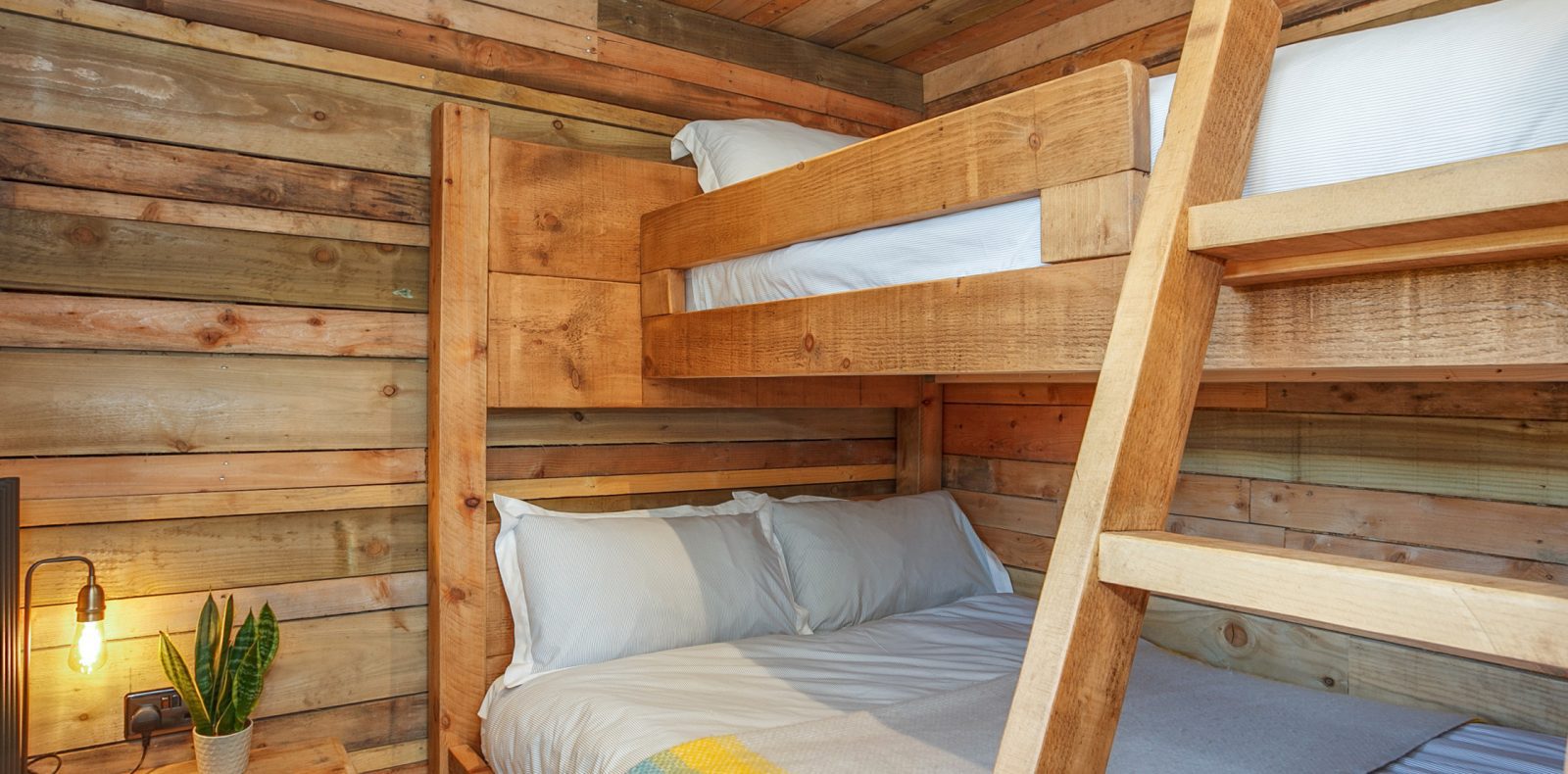 A cozy wooden cabin bedroom, reminiscent of Prince Llywelyn's treehouse, features bunk beds made of rustic planks. The lower bed is neatly made with gray and white bedding. A wooden ladder leads to the upper bunk, adorned with white bedding. Beside the lower bed, a small potted plant and wall lamp add charm.