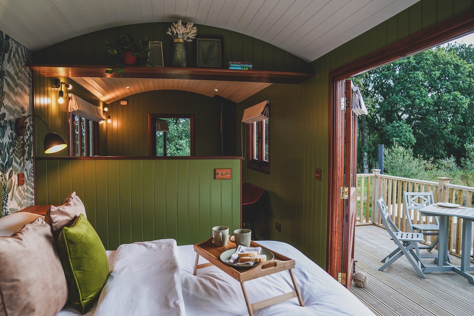 A cozy, green-painted cabin with a bed, topped with white linens and green cushions. A tray with breakfast items is on the bed. The cabin has wooden accents, warm lighting, and a large open door leading to a deck set amidst lush greenery. For an escape like this, list with us today.