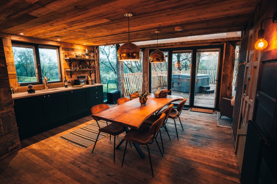 A cozy, rustic dining room with wooden walls, ceiling, and floor. A large wooden table with six chairs is centered under copper pendant lights. Reminiscent of a treehouse, the room has large windows and a glass door opening to a patio with a scenic view of trees and nature outside.