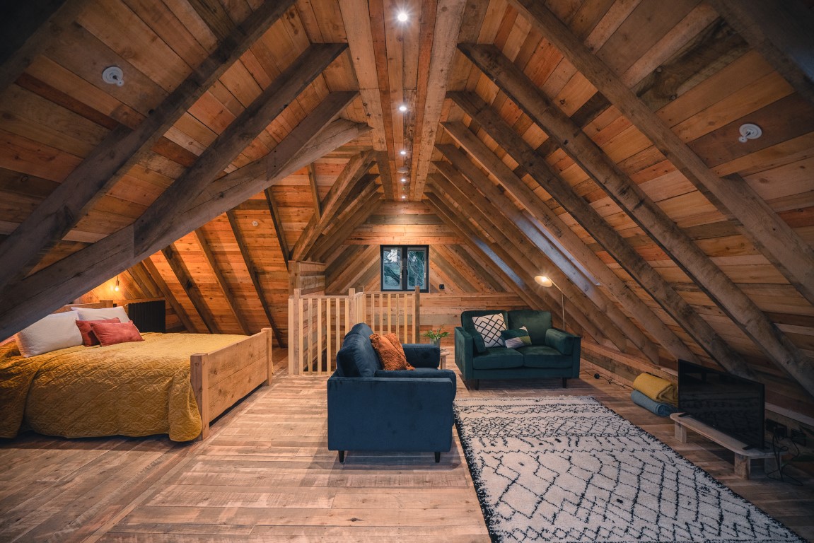 A cozy wooden attic room reminiscent of a treehouse with a large bed on the left featuring yellow bedding. There’s a sitting area in the center with two blue velvet sofas, a rug, and a small TV. The peaked ceiling has exposed wooden beams, and a window provides natural light at the far end.