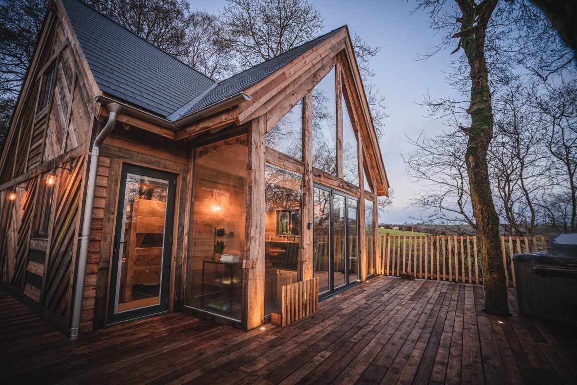 A wooden cabin with large glass windows and a slanted roof sits on a raised treehouse-style deck surrounded by trees. It is evening, with lights illuminating the cozy interior. A fence encloses the area, and a rolling landscape reminiscent of Prince Llywelyn's era is visible in the background.