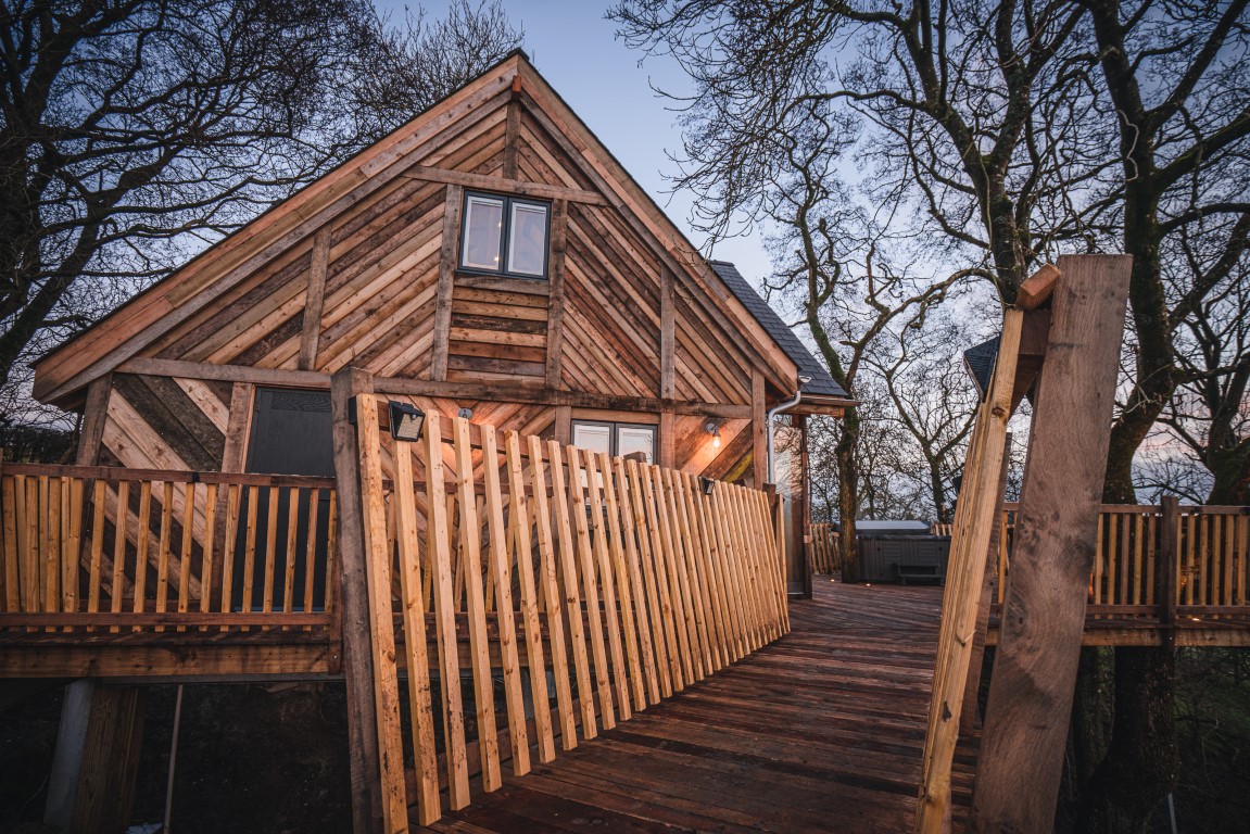 A charming wooden cabin on stilts, reminiscent of Prince Llywelyn’s times, is connected by a wooden bridge. The treehouse features rustic diagonal wood plank siding and a steeply pitched roof. Bare trees surround the structure, and an outdoor area with railings and warm lighting is visible.