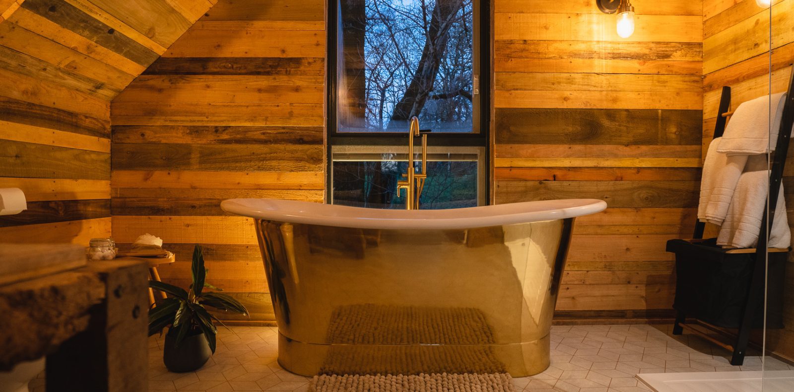 A warmly lit rustic bathroom, reminiscent of Prince Llywelyn's era, features a gold freestanding bathtub in the center, surrounded by wooden plank walls. A black-framed window above the tub showcases a view of bare trees. Towels hang on a black ladder rack to the right, and a potted plant sits near the bathtub.