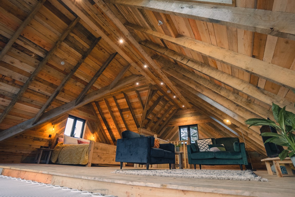 A cozy, rustic attic living space with wooden walls and ceiling beams gives off a charming treehouse vibe. The room features green and dark blue armchairs, a coffee table, and a bed with a yellow blanket. Warmly lit with recessed lighting and a floor lamp, it also boasts a potted plant in the corner.