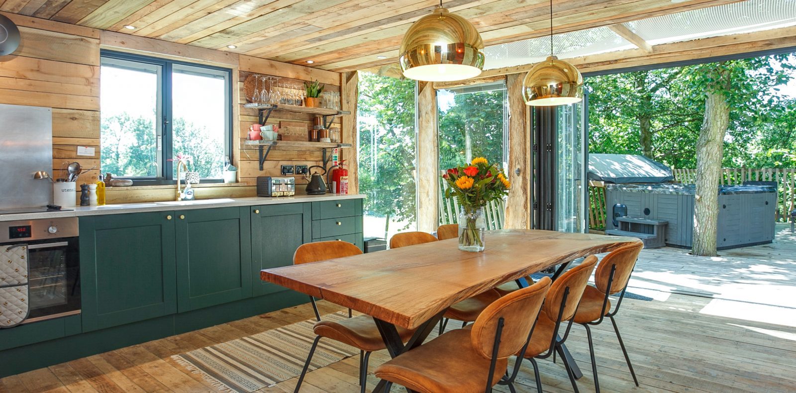 A rustic treehouse kitchen and dining area with wooden walls and floors. The kitchen features green cabinets, modern appliances, and a large wooden table with six chairs. Pendant lights hang above the table, and a vase of flowers sits in the center. Large windows and a sliding door overlook a deck with a hot tub among trees.