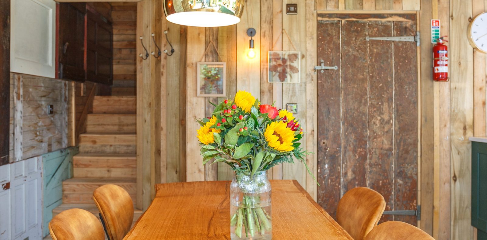 A cozy rustic kitchen with wooden walls features a long wooden dining table adorned with a glass vase of colorful sunflowers. Reminiscent of Prince Llywelyn's treehouse, the room is lit by a warm hanging light, and in the background, there are wooden stairs, framed pictures, a clock, and a fire extinguisher.