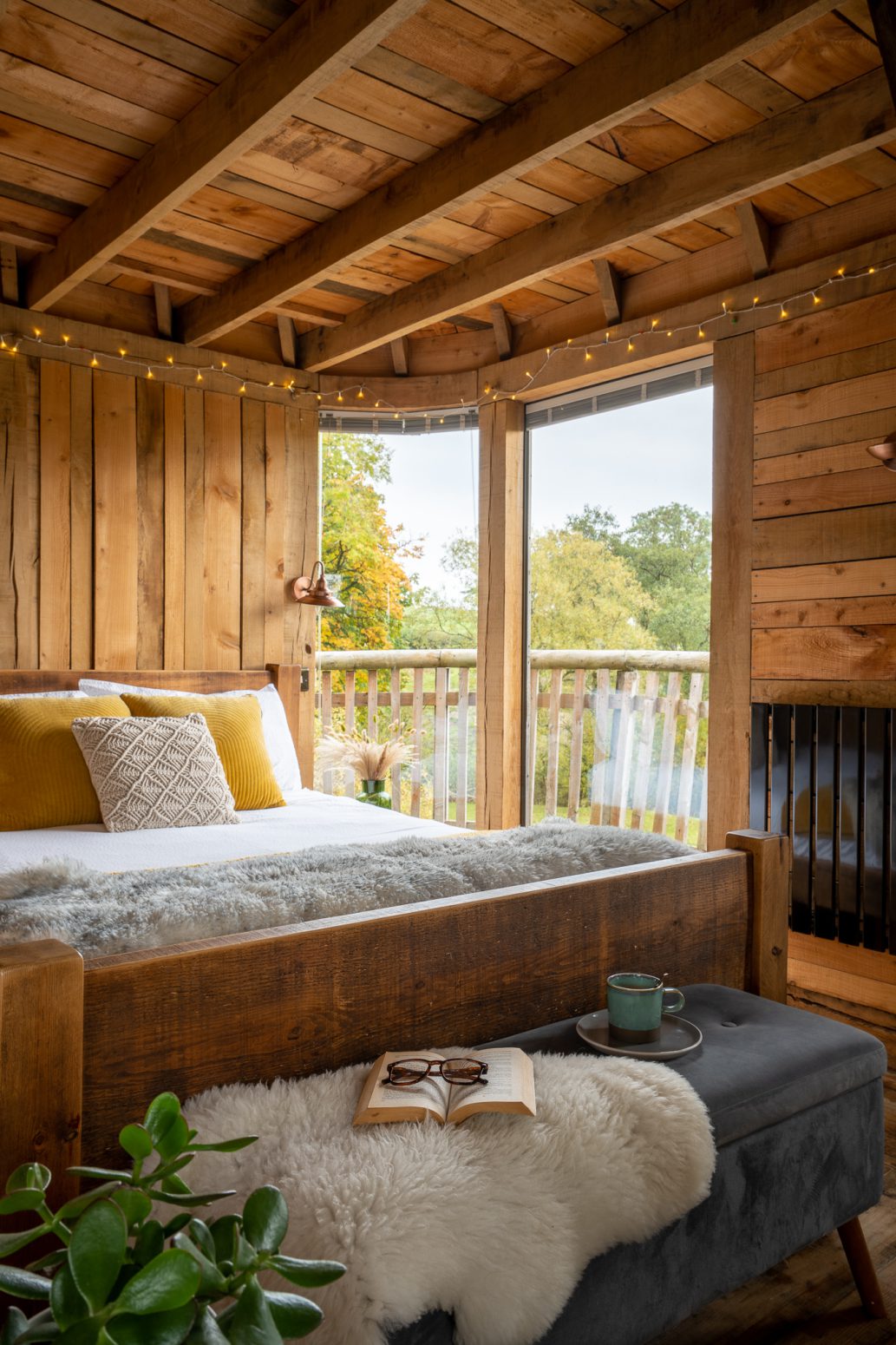 A cozy wooden bedroom with a large bed adorned with yellow and white pillows. The room features fairy lights, a soft blanket, a lush green plant, and a book with glasses on an adjacent bench. Large windows offer a view of greenery and let in natural light, creating the perfect page out of a dream home.