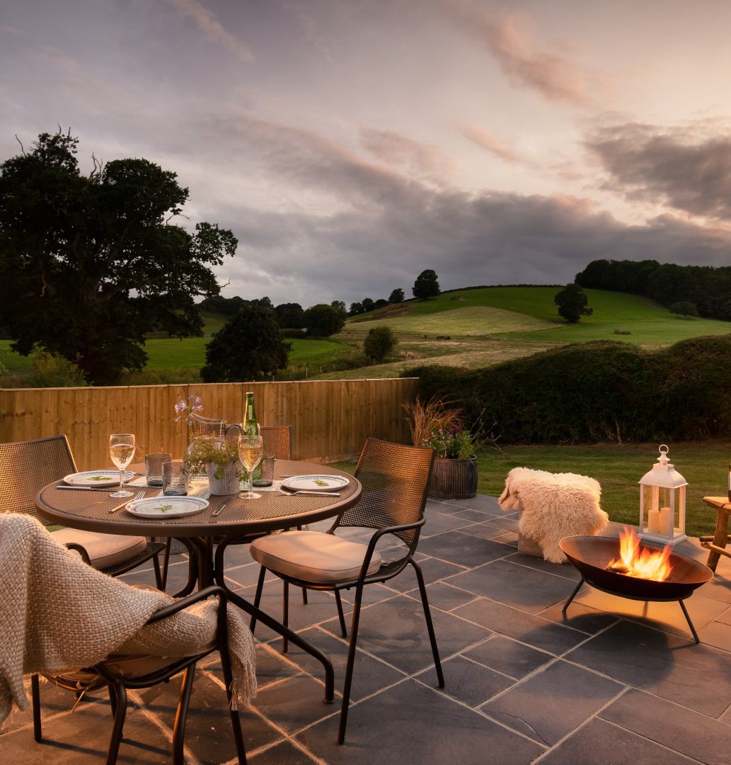 A serene outdoor patio set with a round table and four chairs adorned with white cushions and blankets. The table is set for a meal, complete with wine glasses and a bottle, perfect for showcasing on your Home Page. A glowing fire pit and lanterns enhance the ambiance, amidst rolling hills at sunset.