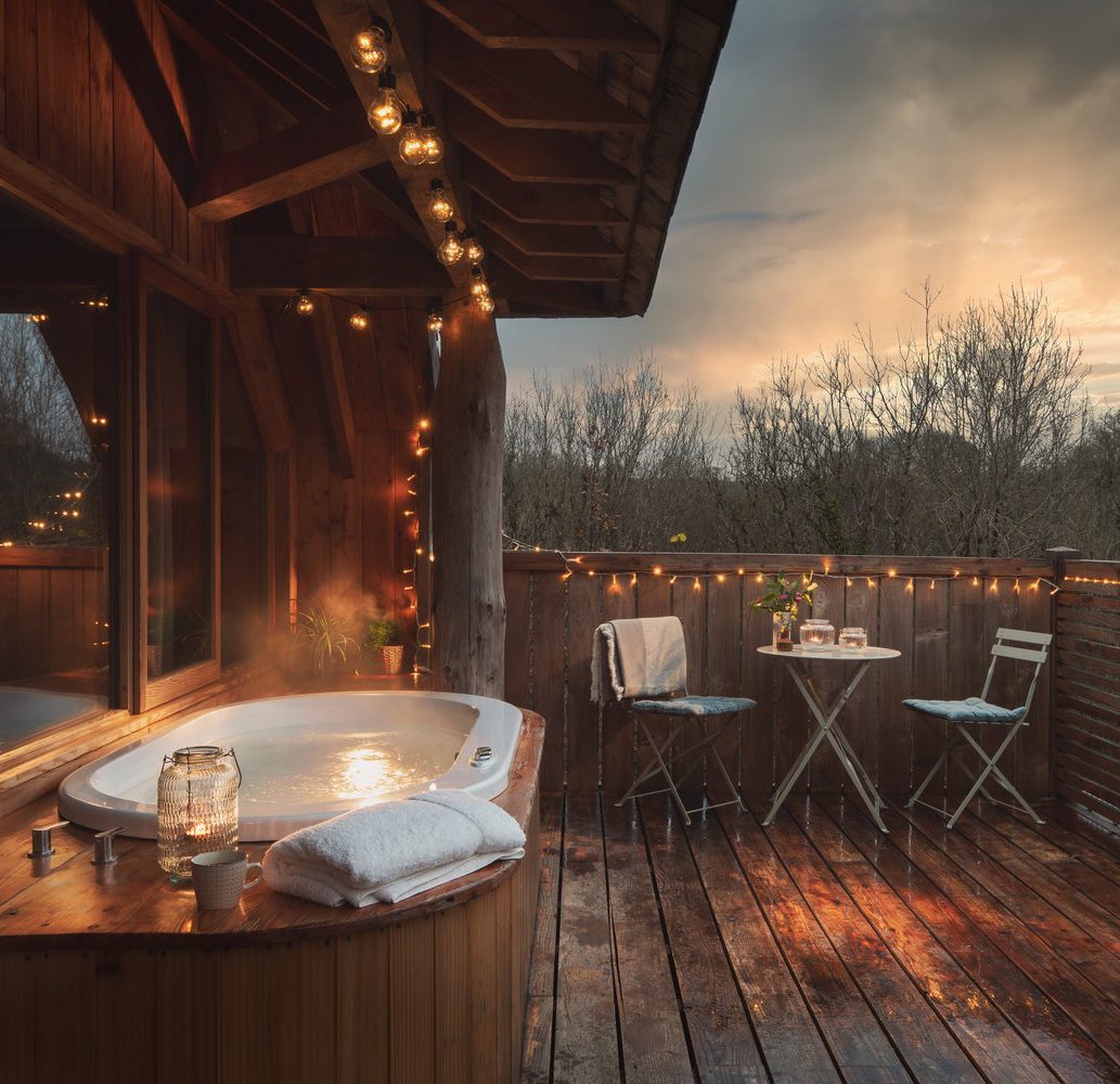 A cozy outdoor wooden deck features a steaming hot tub, surrounded by warm string lights. A small table with two chairs is set up with a beverage and dessert. The background shows bare trees under a dramatic, cloudy sky with rays of sunlight piercing through, creating a perfect home page-worthy setting.