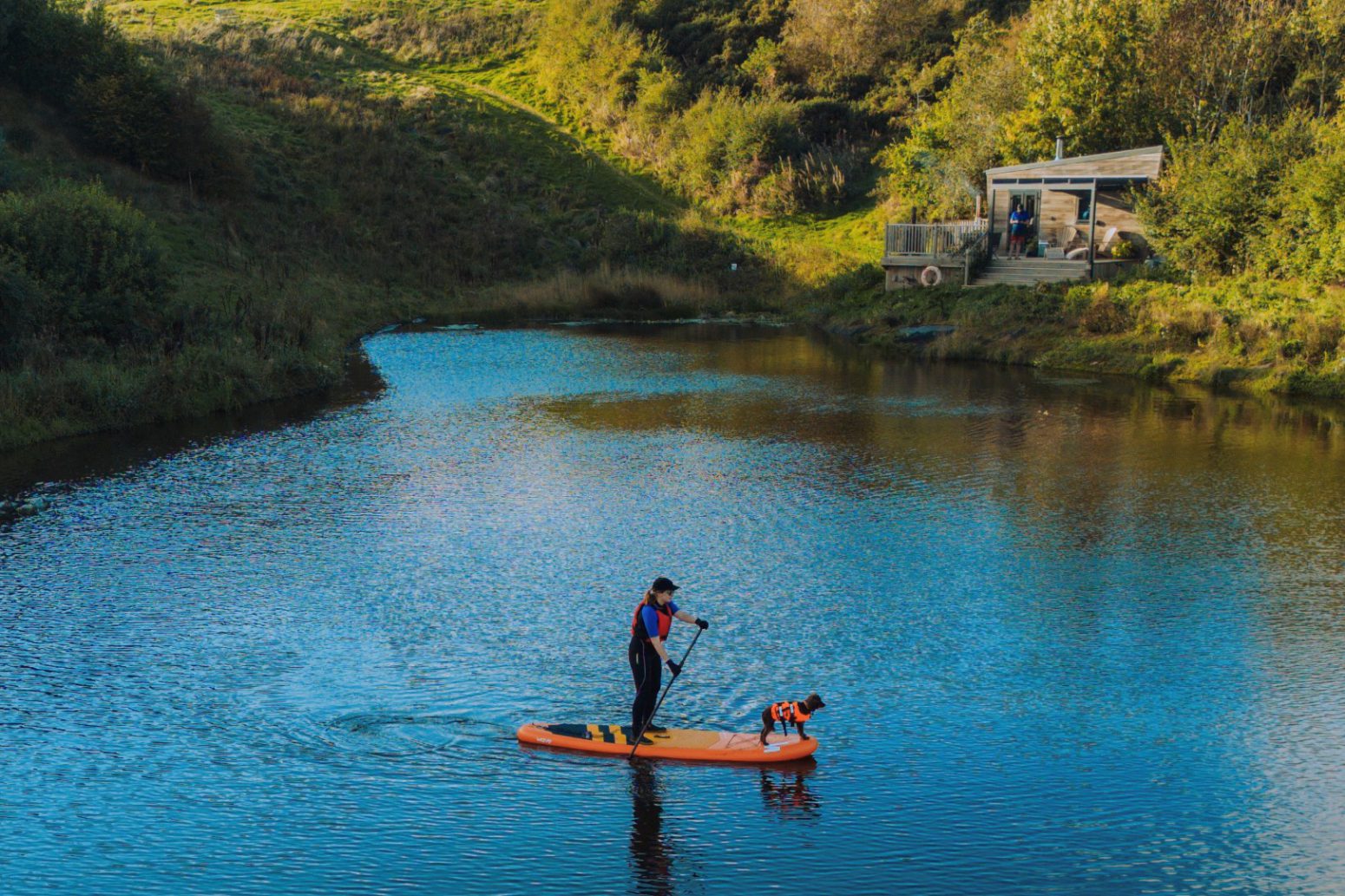 A person stands on an orange paddleboard on a tranquil river surrounded by greenery, wearing a life jacket and a hat. A dog, also wearing a life jacket, stands at the front of the paddleboard, looking at the water. A small cabin is visible in the background, like something straight from your Home Page.