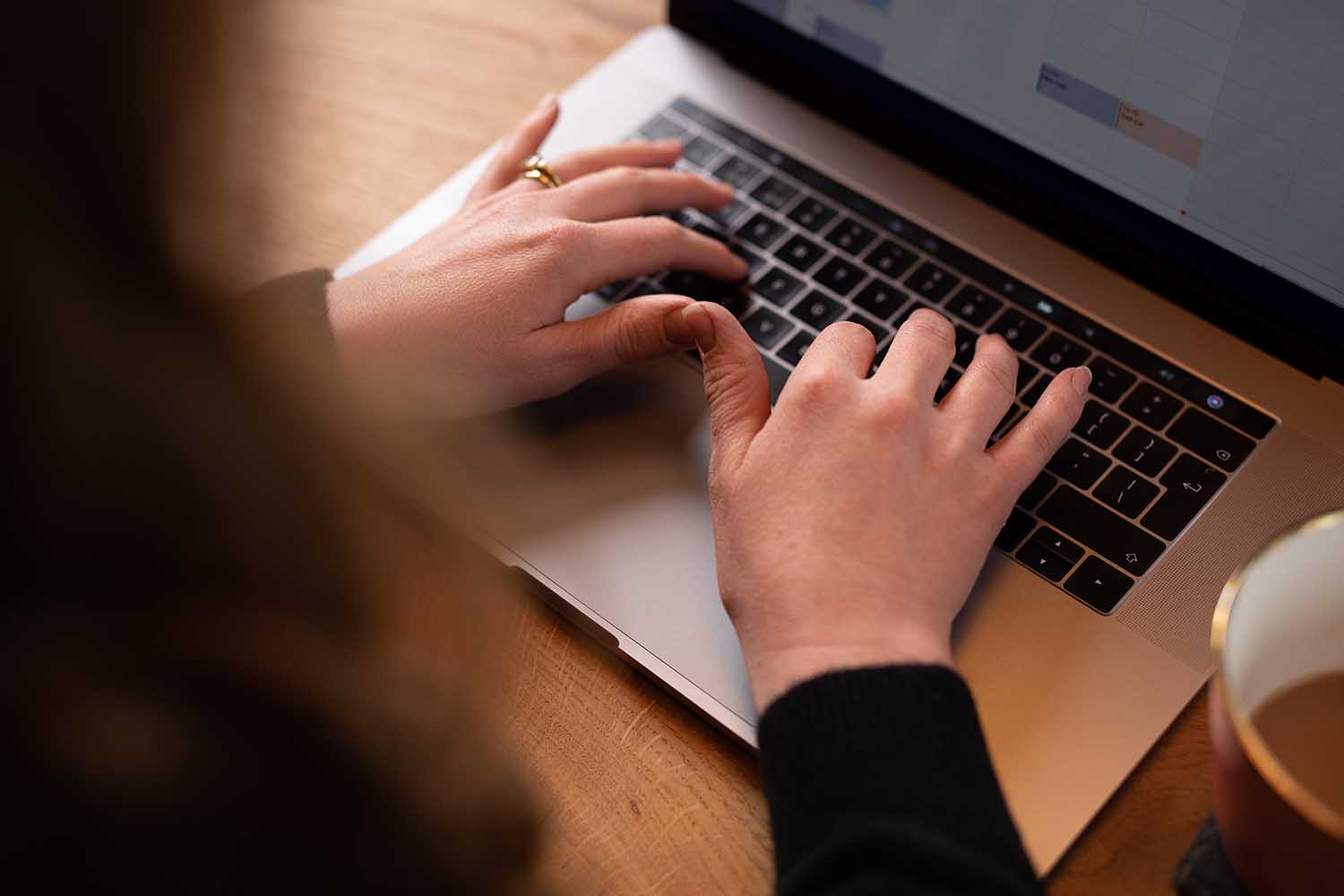 A person types on a laptop keyboard, with a partially visible screen displaying a calendar—likely planning their next post for ToWander travel blog. The individual wears a black sleeve and a gold ring on the right hand. A wooden surface and the edge of a coffee mug are visible in the background.