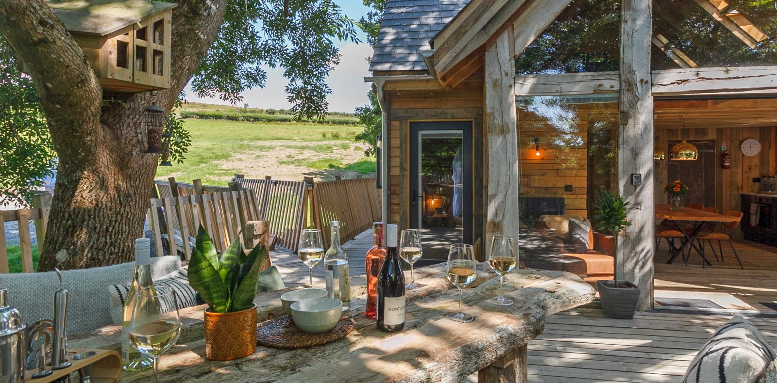 A rustic outdoor patio with a long wooden table set with wine bottles, glasses, and a potted plant. The enchanting Prince Llywelyn Treehouse is built around a nearby tree. A cozy cabin with large glass windows overlooks an idyllic countryside with green fields in the background.