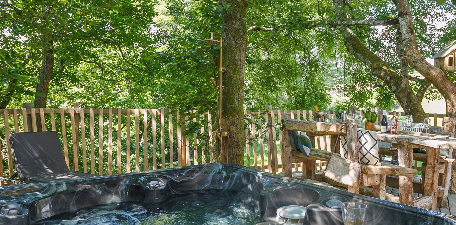 A wooden deck surrounded by lush greenery features a hot tub in the foreground, a rustic wooden table with drinks and snacks, and two lounge chairs. A wooden fence encloses the area, with a small treehouse visible among the trees, creating an enchanting retreat fit for Prince Llywelyn.