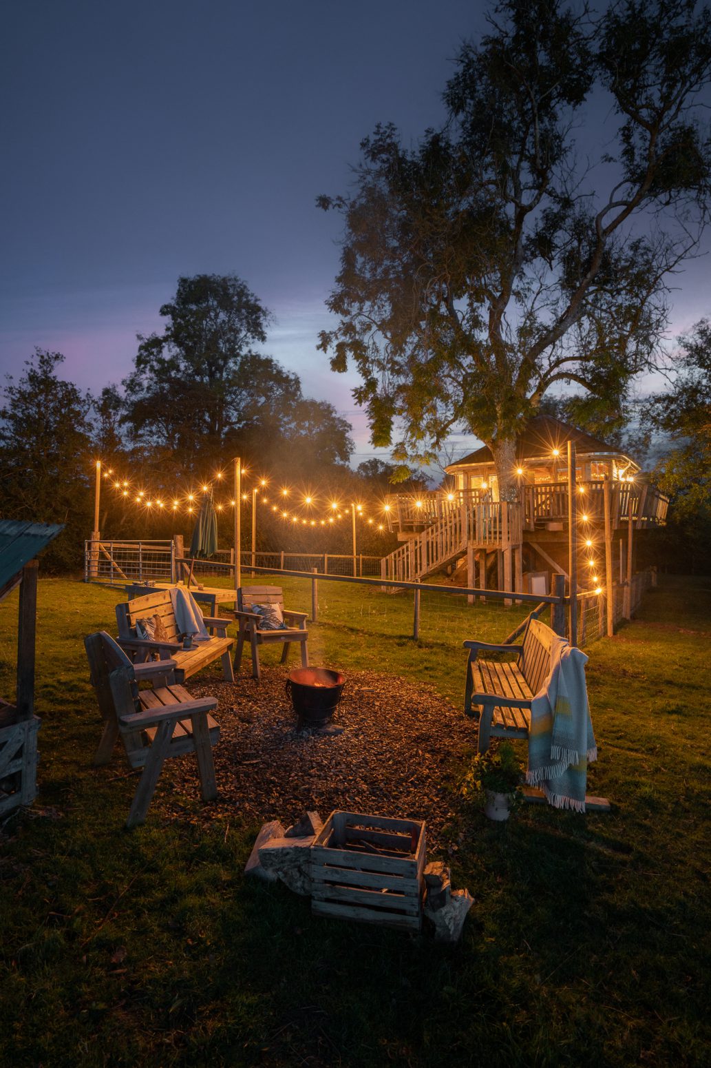 A cozy outdoor seating area is shown at dusk, with wooden benches arranged around a fire pit. String lights hang above, illuminating the scene. In the background, a treehouse with a ladder and railing is visible, surrounded by tall trees and a partly cloudy sky. It feels like home.