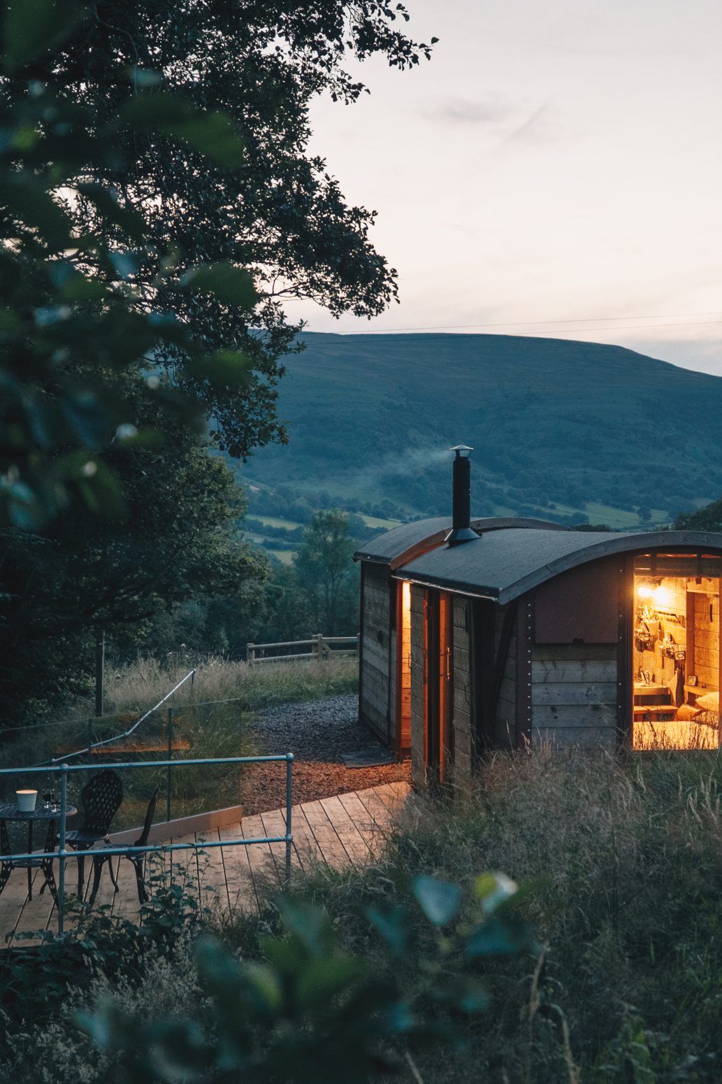 A cozy, illuminated tiny house nestled among tall grass and trees at dusk. The structure overlooks a scenic landscape with rolling hills and mountains in the background. A wooden deck with chairs is visible outside the house, surrounded by lush greenery—a perfect feature for your website's home page.