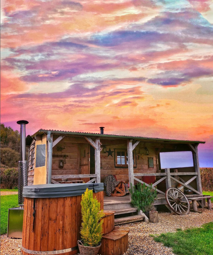 A rustic wooden cabin stands under a vibrant, colorful sunset sky with hues of pink, purple, and orange. In front of the cabin, there is a circular wooden hot tub, a barrel, and various plants. A wagon wheel is also visible near the entrance. This charming getaway awaits—list with us for an unforgettable experience!