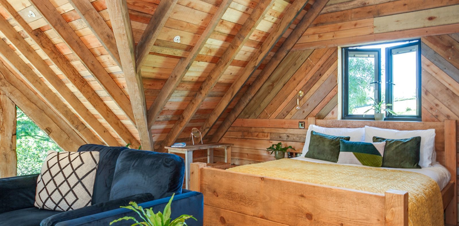 A cozy Prince Llywelyn Treehouse loft with wooden walls and ceiling. The space features a bed with green and yellow pillows, a yellow blanket, and a small wooden nightstand. A navy blue couch with a patterned cushion sits nearby, and a window with a view of trees is open.