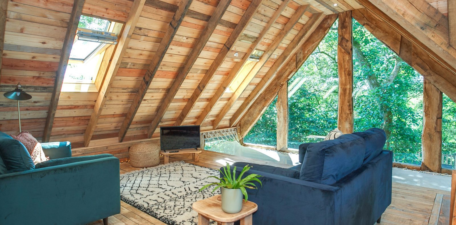 A cozy attic living room with wooden walls and a pitched roof, reminiscent of a rustic treehouse. It features a large window with a view of lush trees, two blue armchairs, a small wooden table with a potted plant, a floor lamp, and even Prince Llywelyn would feel at home here.