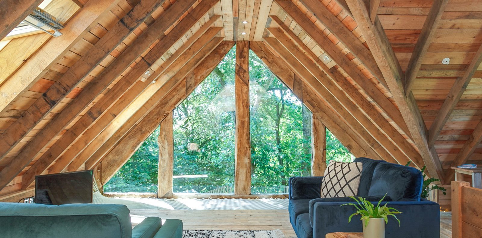 A cozy triangular attic space with wooden beams and large floor-to-ceiling windows overlooking lush greenery, reminiscent of the Prince Llywelyn Treehouse. The room features comfortable seating, including a blue armchair and a gray sofa, and a small wooden coffee table with plants.