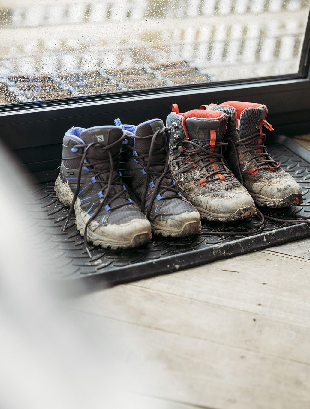 Two pairs of muddy hiking boots, one with blue laces and the other with red laces, are placed side by side on a black mat near a glass door. The door overlooks a rainy scene outside. Perfect for our next ToWander travel blog entry!