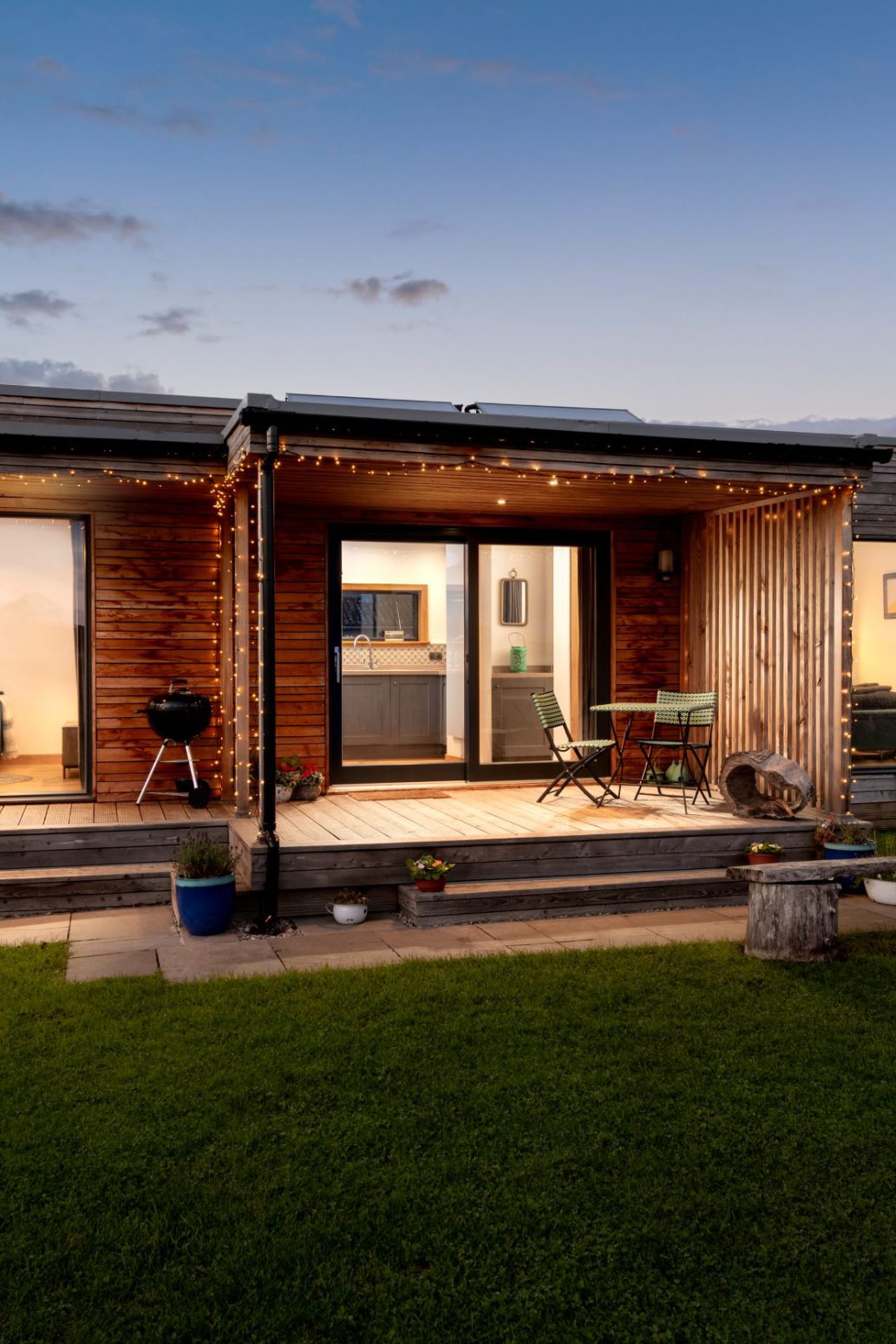 A modern, single-story wooden house with large glass doors and windows, illuminated by string lights at dusk. The house boasts an outdoor seating area with a grill and various plants. In the foreground, there's a lit fire pit on a grassy lawn—a perfect spot for those who travel in search of adventure.