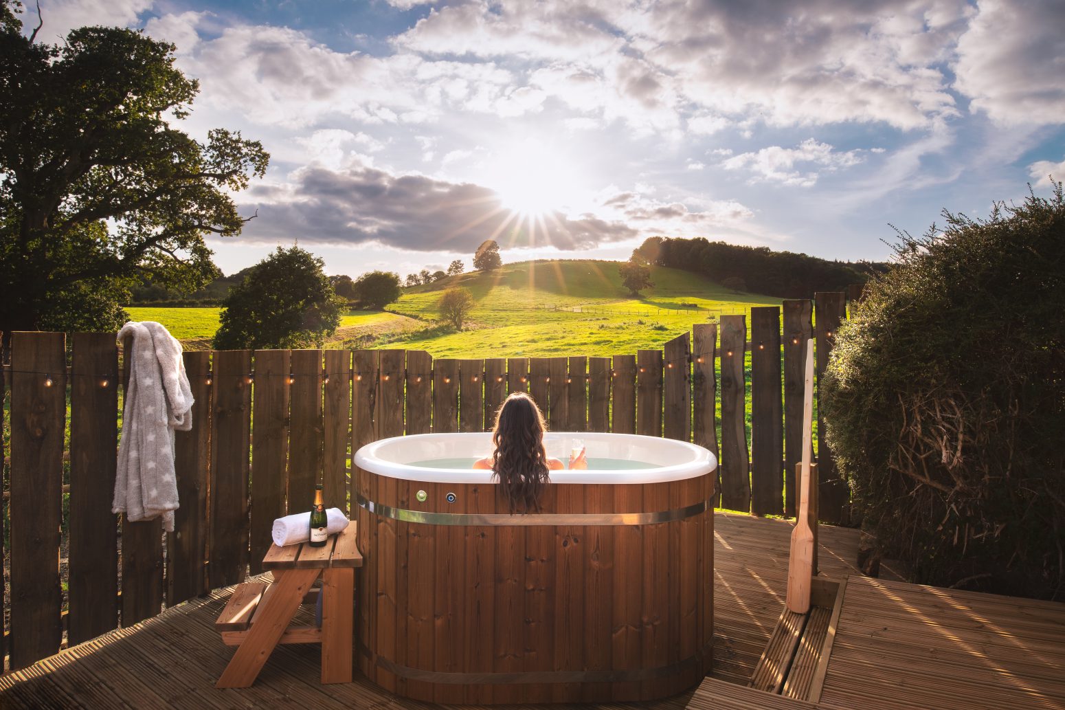 A person with long hair relaxes in an outdoor wooden hot tub, facing a scenic view of green fields and a setting sun. The hot tub is surrounded by a wooden fence, with a robe and towel hanging nearby and a bottle of wine on a small wooden table—an ideal spot for those who love wandering activities.