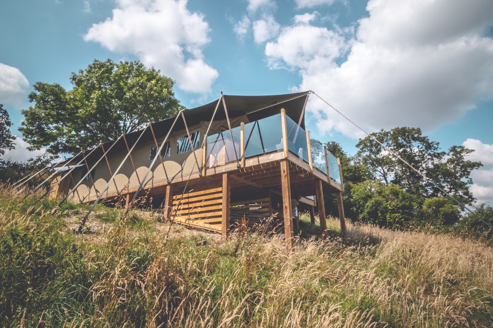 A modern, elevated tent-like home in a grassy, rural setting. The building features large glass panels and canvas coverings supported by poles. It's surrounded by tall grass and trees, with a partly cloudy sky as the backdrop.