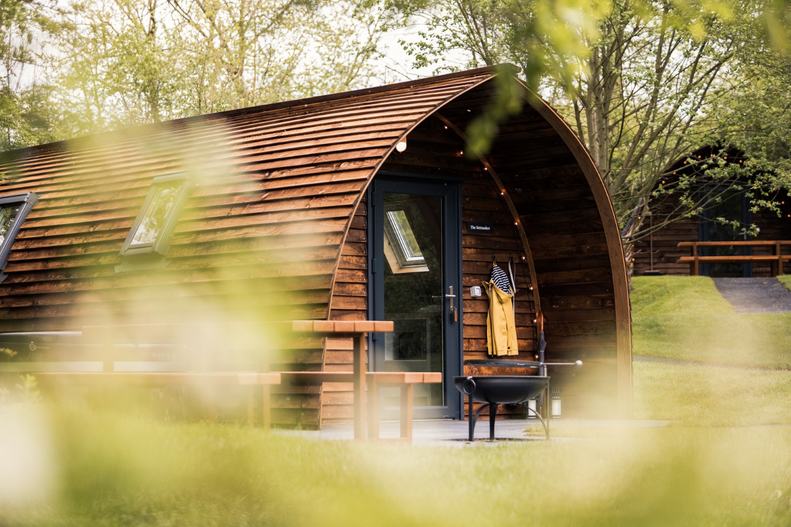 A cozy, wooden cabin with a curved roof nestled in a lush, green outdoor setting. The home has a small porch with a chair and a jacket hanging near the entrance. Sunlight filters through surrounding trees, creating a peaceful, natural atmosphere.