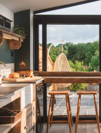 A bright home kitchen with a wooden countertop and stools, featuring a cutting board with bread and fruits. Through the large sliding glass door, a serene patio with a hanging chair and lush green trees is visible. The space exudes a warm and inviting atmosphere, perfect for any page in your life.