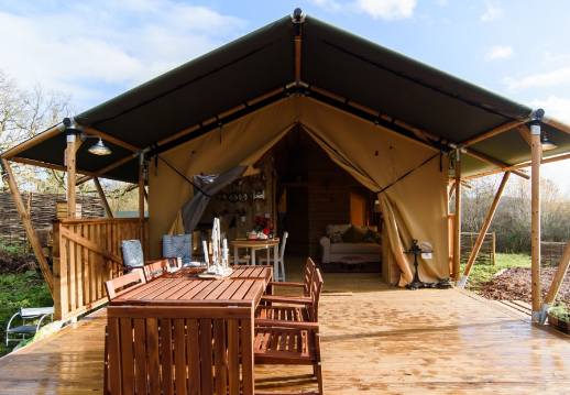 A glamping tent with an open front stands on a wooden deck, creating a picturesque home away from home. The deck features a dining table with six chairs. Inside the tent, there are a cozy bed and a small dining table, surrounded by lush greenery and trees under a partly cloudy sky—right out of a storybook page.