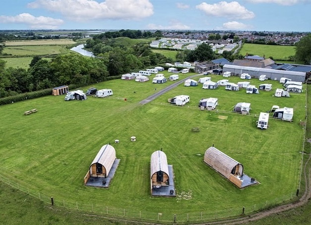 Aerial view of a green camping site at Abbey Farm, where several white caravans and motorhomes are parked around the area. Three wooden glamping pods are arranged in a row in the foreground, with a river and scattered buildings visible in the background.