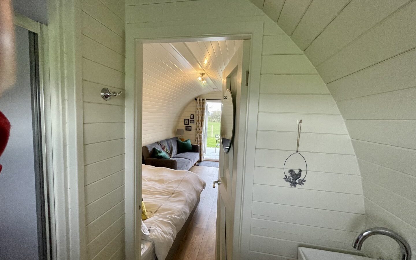 A cozy, small cabin interior at Abbey Farm with white-paneled walls and ceiling. The view is through a doorway showing a bed on the left, a couch and small table against the far wall with a window overlooking greenery. A small sink is visible on the right—perfect for glamping enthusiasts.