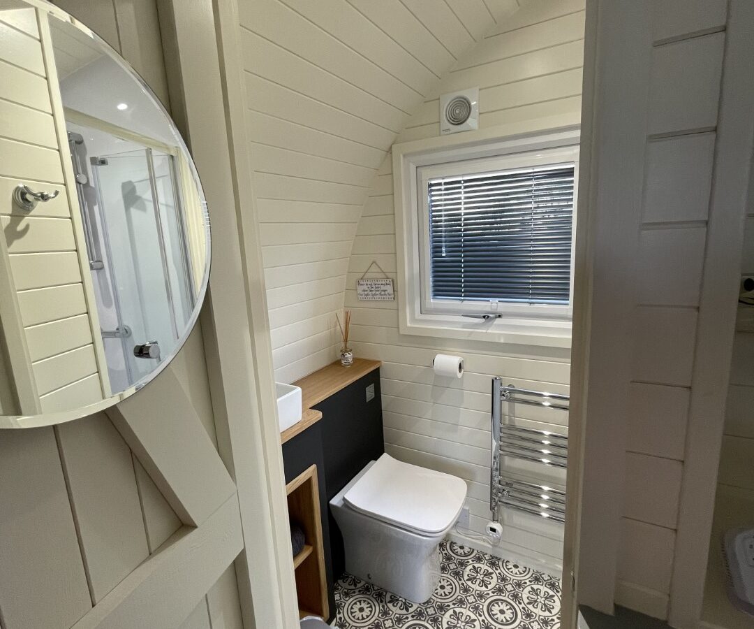 A small, cozy bathroom with white wooden paneling and patterned floor tiles, perfect for your glamping retreat at Abbey Farm. It features a wall-mounted toilet, a round mirror on the open door, a window with blinds, silver towel rack, toilet paper holder, trash can and toiletries shelf.